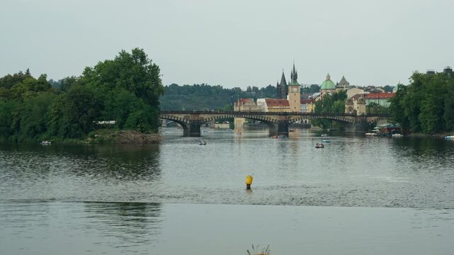 People ride pedal boats on Vltava River in Prague. Historic Charles Bridge stands in background. Scenic urban panorama depicts summer travel, European vacation, and water leisure activity.