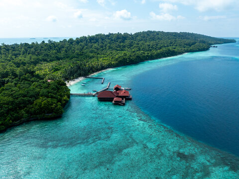 Aerial view of a tropical island resort with wooden overwater bungalows surrounded by turquoise coral reef waters and lush green forest in Berau Regency, East Kalimantan, Indonesia.