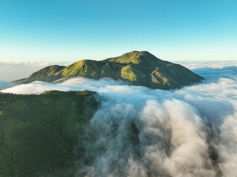 Aerial view of Gunung Mongkrang rising above a thick sea of clouds with lush green slopes under a clear blue sky in Jatiyoso, Central Java, Indonesia.