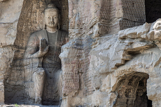 View of the large carved Buddha statue at the Yungang Grottoes, featuring intricate stone carvings and smaller figures on the surrounding rock walls in Datong, Shanxi, China.