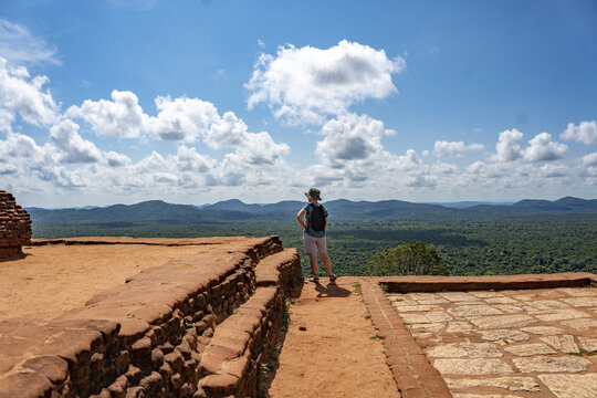 Sigiriya, Sri Lanka - 26 April 2026: View of a tourist standing on the historic Sigiriya Lion Rock, overlooking the vast green jungle landscape under a bright blue sky with fluffy white clouds.