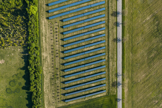 Aerial view of a solar farm with rows of photovoltaic panels arranged in a field next to a road, surrounded by green meadows and trees in Kunkovice, Zlin Region, Czechia.