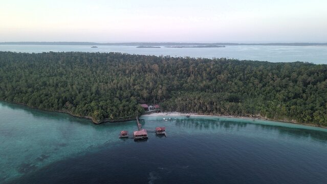 Aerial view of wooden overwater bungalows on stilts connected by a pier to a lush tropical island with dense forest and white sand beaches, Pulau Derawan, East Kalimantan, Indonesia.