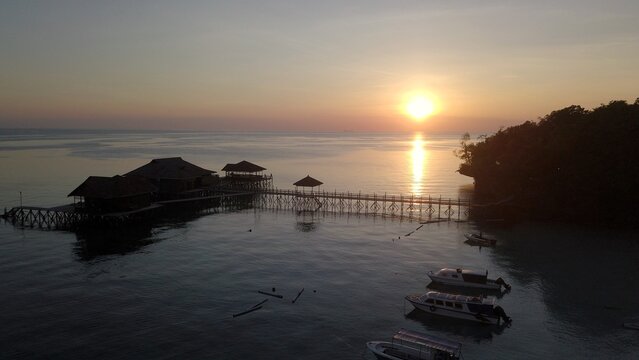 Aerial view of wooden overwater bungalows and a long pier extending into the calm sea at sunset with moored boats in Pulau Derawan, East Kalimantan, Indonesia.