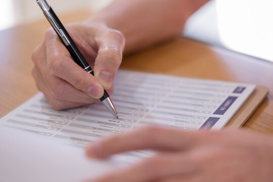 Person completing a questionnaire with pen on paper, representing survey, assessment, feedback, evaluation, checklist, research, data collection and customer satisfaction in a professional setting.