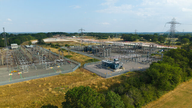 Aerial view of Berkswell Substation with electrical transformers, high voltage power lines, and metal fencing surrounded by green fields and trees in Coventry, England, United Kingdom.
