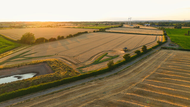 Aerial view of a combine harvester working in golden wheat fields during sunset with wind turbines in the distance and a small pond in Enderby, England, United Kingdom.