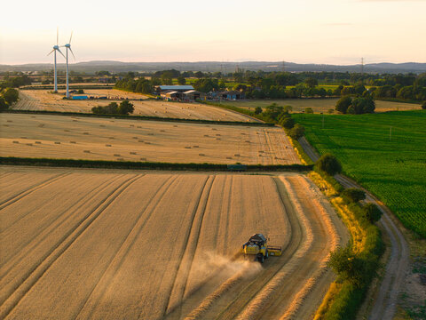 Aerial view of a combine harvester working in golden wheat fields during harvest season with wind turbines in the background near Enderby, England, United Kingdom.