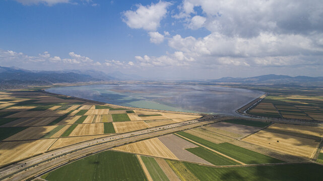 Aerial view of Sugla Lake surrounded by vast agricultural fields and patchwork farmlands under a bright blue sky with scattered clouds in Ahirli, Konya, Turkiye.