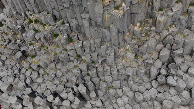 Aerial view of geometric basalt columns forming a rugged volcanic rock wall with moss patches in Iceland.