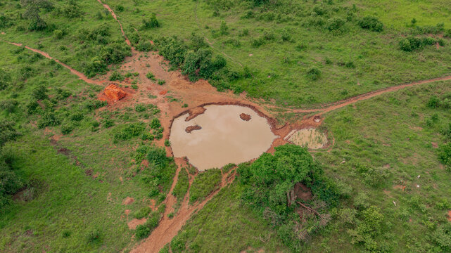 Aerial view of a small muddy watering hole surrounded by lush green vegetation, dirt paths, and scattered trees in the rural landscape of Kapelebyong, Eastern Region, Uganda.