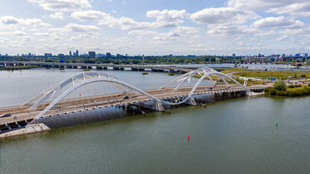 Aerial view of the Enneus Heermabrug, a modern white arched bridge spanning the water with a distant city skyline under a blue sky with clouds in Amsterdam, Noord-Holland, Netherlands.
