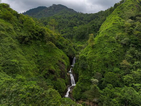 Aerial view of Curug Jagapati waterfall cascading through lush green tropical forest and steep hills in Cisompet, West Java, Indonesia.