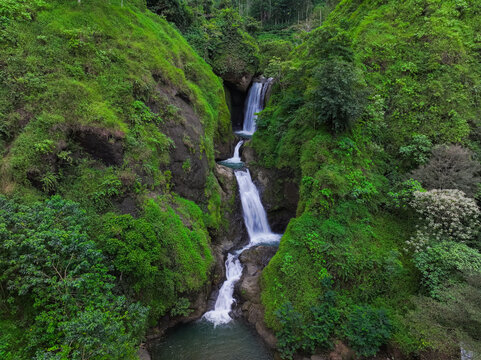 Aerial view of Curug Jagapati waterfall cascading down rocky cliffs surrounded by lush green tropical vegetation in Cisompet, West Java, Indonesia.