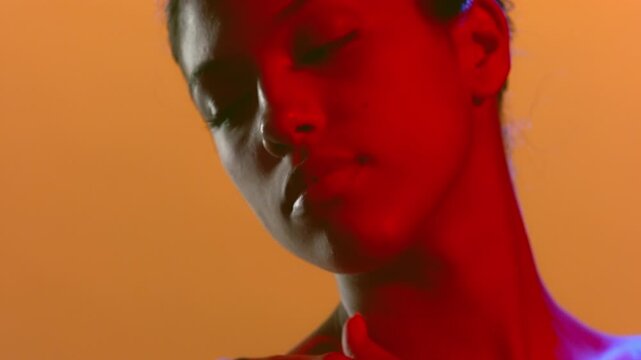 Close-up shot of sensual beauty model lit with blue and red led touching her nape and turning her head looking aside and down set against an orange bg | Fragrance campaign footage
