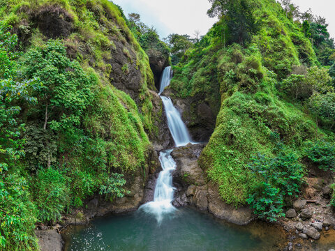 Aerial view of Curug Jagapati waterfall cascading down a lush, green, moss-covered rocky cliff into a clear pool in Cisompet, West Java, Indonesia.