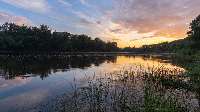 A serene river flows gently at sunset, its calm surface mirroring the vibrant orange clouds and dense, dark green trees
