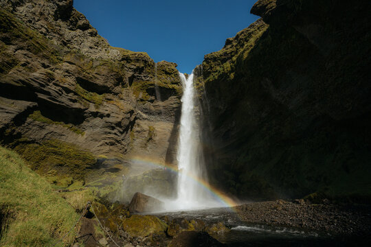 View of Kvernufoss waterfall cascading down a rocky canyon with a rainbow at the base, surrounded by mossy cliffs under a clear blue sky in Evindarholar, Rangarthing eystra, Iceland.