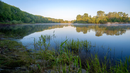 Obraz premium A calm river reflects lush green trees and a clear blue sky on a tranquil, bright and sunny morning.
