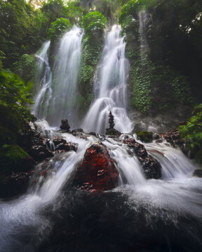 View of a cascading waterfall surrounded by lush tropical greenery, moss-covered rocks, and stacked stone cairns in Bali, Indonesia.