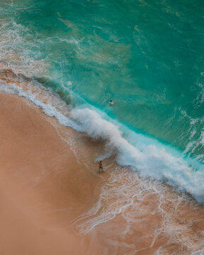 View of Kelingking Beach with turquoise waves crashing onto the sandy shore and people swimming and walking in Nusa Penida, Bali, Indonesia.