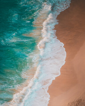 View of the turquoise waves crashing onto the golden sands of Kelingking Beach in Nusa Penida, Bali, Indonesia.