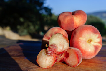 Sweet red flat peaches, one sliced open, showing the juicy flesh and small pit, sitting on a wooden surface in summer light © JOSEANTONA