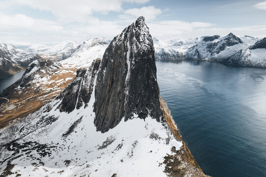 Aerial view of the dramatic Segla mountain peak rising above the deep blue fjord, surrounded by snow-covered slopes and rugged arctic landscape in Senja, Troms, Norway.