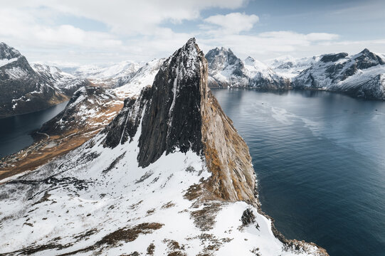 Aerial view of Segla mountain rising above the deep blue waters of the fjord, surrounded by snow-capped peaks and rugged coastal landscape in Senja, Troms, Norway.