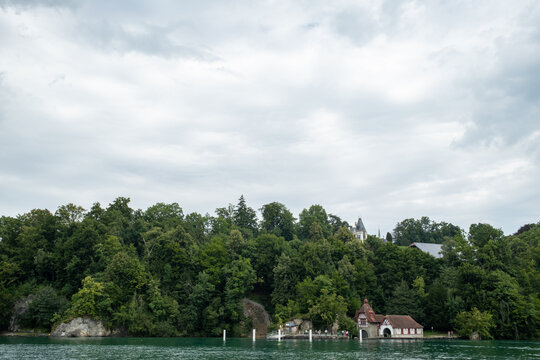 View of a historic building nestled among dense green trees on the rocky shoreline of Lake Lucerne, Lucerne, Lucerne, Switzerland.