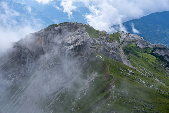 View of Mount Pilatus with a paraglider soaring over the rocky mountain ridge and green slopes partially covered by clouds in Alpnach, Obwalden, Switzerland.