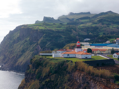 Aerial view of Lajes das Flores lighthouse on a dramatic cliffside overlooking the Atlantic Ocean, with lush green hills and coastal vegetation in Lajes das Flores, Azores, Portugal.