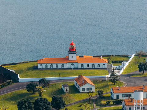 Aerial view of the Lajes das Flores Lighthouse with its red lantern and orange roof, set on a green coastal cliff overlooking the Atlantic Ocean in Lajes das Flores, Azores, Portugal.