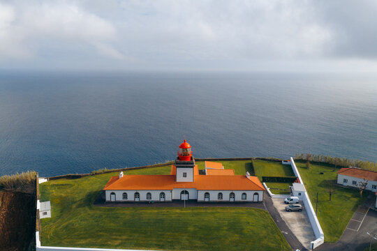 Aerial view of the Lajes das Flores Lighthouse with its red lantern room and orange roof, set on a grassy cliff overlooking the Atlantic Ocean in Lajes das Flores, Azores, Portugal.