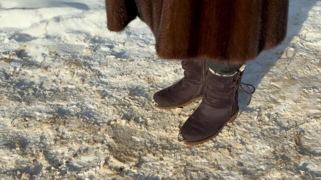 Mink coat suede boots in snow. Woman shifting weight, stomping feet to warm, visible breath in frosty air, sunlight highlighting