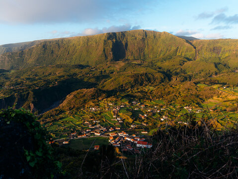 View of the village of Flores Island nestled in a lush green valley with terraced fields and a dramatic mountain backdrop under warm golden hour light in Azores, Portugal.