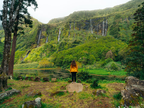 View of a woman in a yellow jacket standing on a rock overlooking the waterfalls of Poco da Alagoinha on Flores Island, Azores, Portugal.