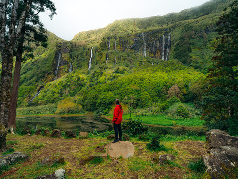 View of a person in a red jacket standing on a rock overlooking the lush green cliffs and multiple waterfalls of Flores Island in the Azores, Portugal.