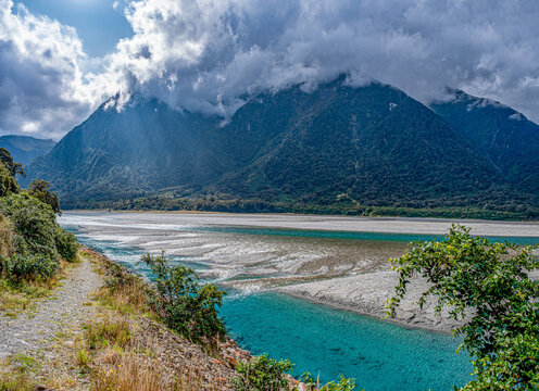 Haast River, Mount Aspiring National Park, breites Flussbett mit tuerkisfarbenem Wasser, Wolken haengen tief ueber bewaldeten Bergen, Wanderweg im Vordergrund, Suedinsel, Neuseeland