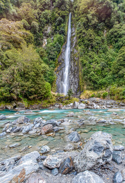 Thunder Creek Falls, Mount Aspiring National Park, Haast Pass, Wasserfall stuerzt durch dichten Regenwald, tuerkisfarbener Fluss mit Felsbrocken im Vordergrund, Suedinsel, Neuseeland