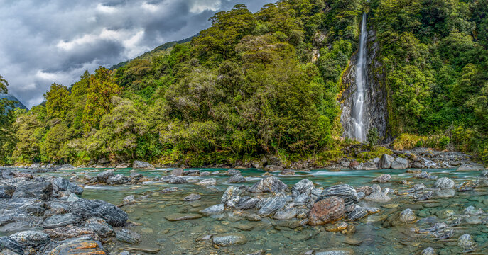 Thunder Creek Falls, Mount Aspiring National Park, Haast Pass, Wasserfall stuerzt durch dichten Regenwald, tuerkisfarbener Fluss mit Felsbrocken im Vordergrund, Suedinsel, Neuseeland