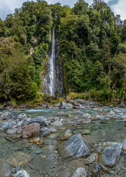 Thunder Creek Falls, Mount Aspiring National Park, Haast Pass, Wasserfall stuerzt durch dichten Regenwald, tuerkisfarbener Fluss mit Felsbrocken im Vordergrund, Suedinsel, Neuseeland
