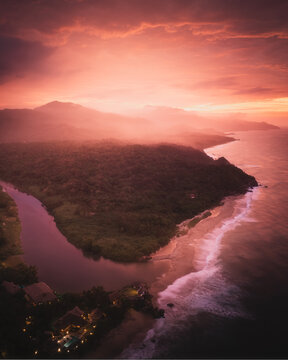 Aerial view of Parque Nacional Natural Tayrona with a river flowing into the sea, lush tropical forest, and mountains under a vibrant sunset in Magdalena, Colombia.