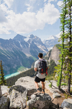 View of a hiker with a backpack on a rocky cliff edge overlooking the turquoise waters of Lake Louise surrounded by snow-capped mountains and pine forests in Lake Louise, Alberta, Canada.
