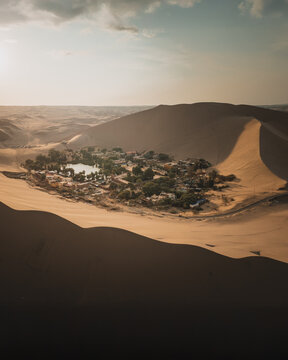 Aerial view of the Huacachina oasis surrounded by vast sand dunes, palm trees, and buildings under a warm sunset sky in Huacachina, Ica, Peru.