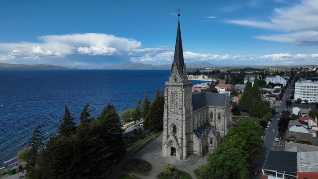 Aerial view of Catedral de San Carlos de Bariloche, a historic stone church with a tall spire on the shores of Nahuel Huapi Lake, surrounded by trees in Bariloche, Rio Negro, Argentina.
