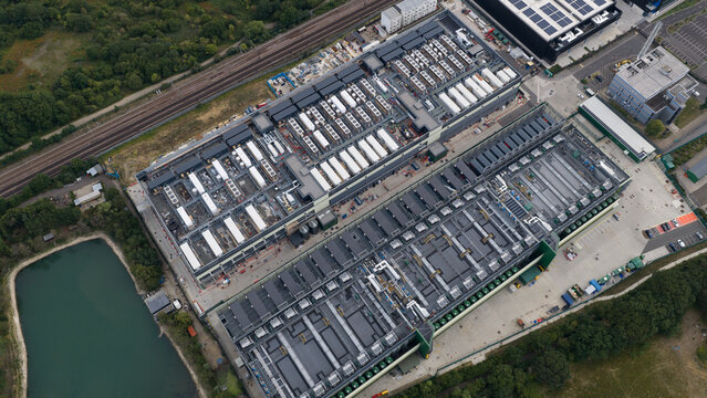 Aerial view of NTT LON1B Data Centre featuring rows of rooftop cooling units and industrial infrastructure situated near a railway line and pond in Dagenham, England, United Kingdom.