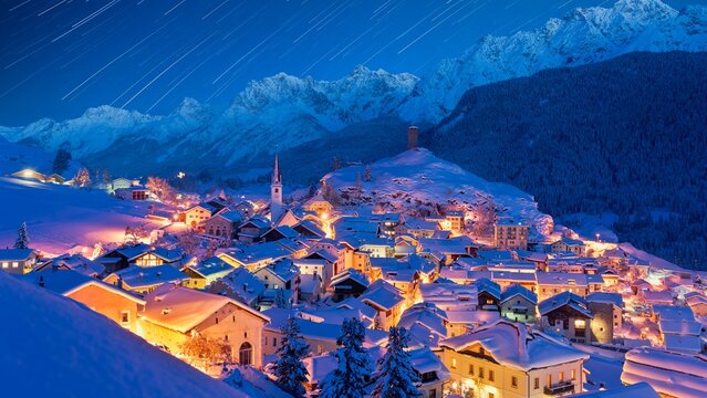 View of the historic village of Ardez at night with snow-covered rooftops, church steeple, and star trails in the sky above the mountains in Scuol, Grisons, Switzerland.