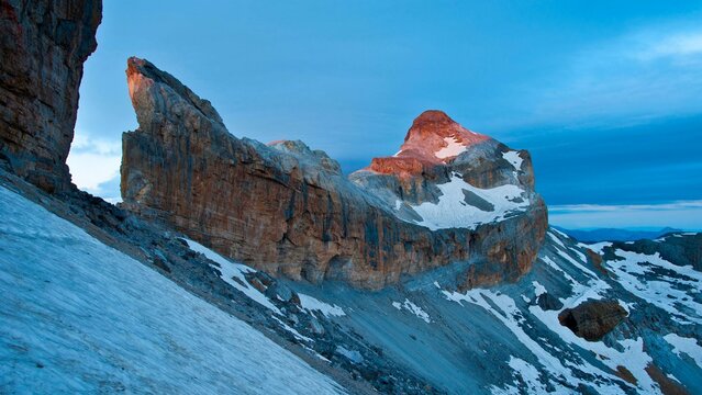 View of La Breche de Roland, a massive natural mountain gap with rugged rocky cliffs and snow-covered slopes illuminated by golden dawn light in Gavarnie-Gedre, Occitanie, France.