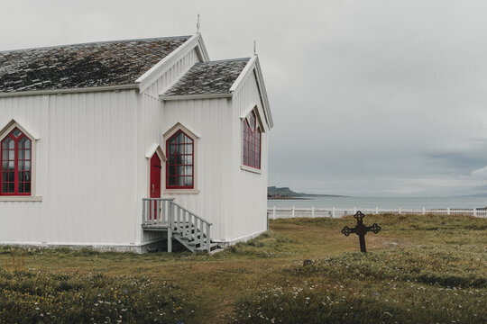 Nesseby, Finnmark, Norway - 26 August 2025: View of the historic white wooden Nesseby Church with red window frames and a small cemetery cross in the foreground under a cloudy sky.